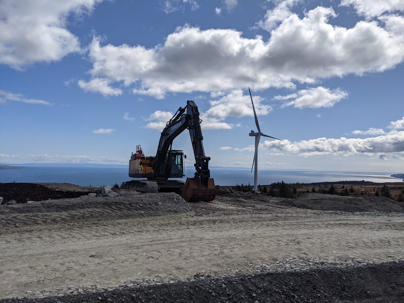 Excavator on a wind farm haul road, with a wind turbine and the sea behind